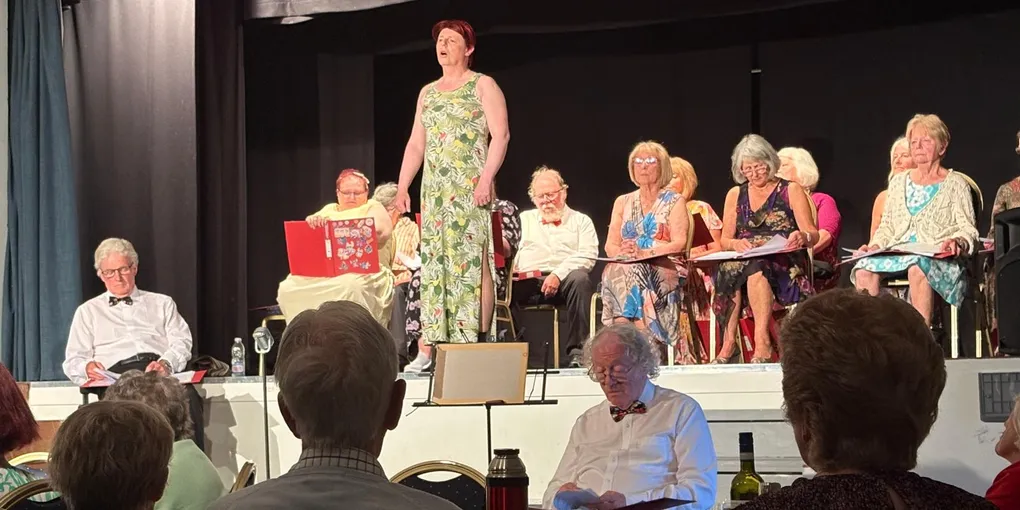 The stage at Lisvane Players memorial hall with performers sitting down. Robyn is standing and singing at the front of the stage. Tables and chairs accommodating audience members are in the foreground.